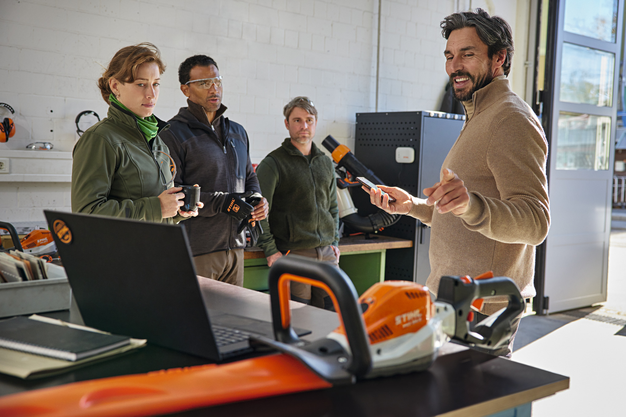Cuatro personas de pie en un taller y una persona explica cómo funciona la aplicación STIHL connected en un smartphone