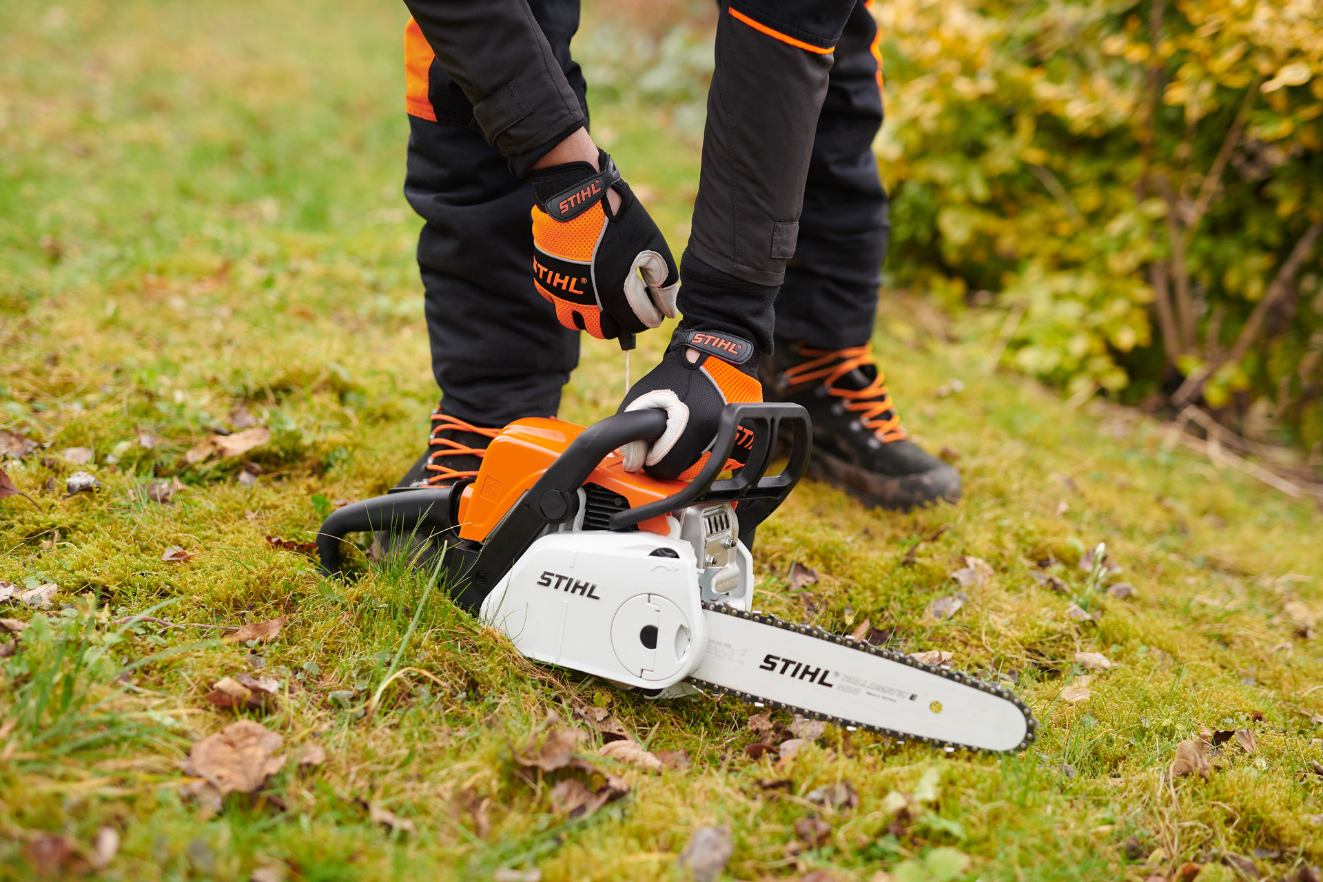 Una motosierra STIHL MS 180 C-BE en el suelo, con una persona que lleva guantes protectores sujetando el asa.