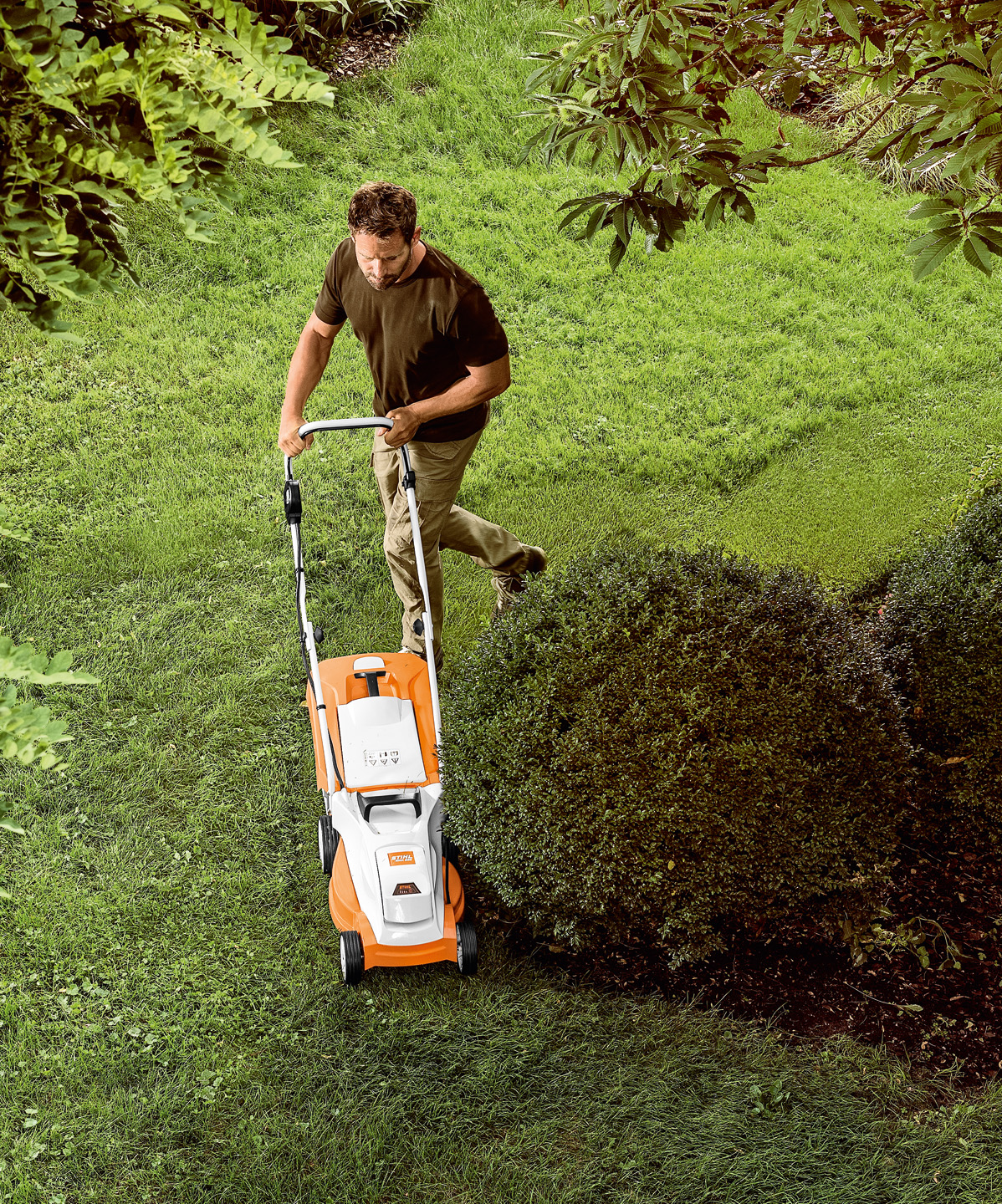 Un hombre corta hierba alrededor de un arbusto con el cortacésped STIHL RMA 235 con batería AK.