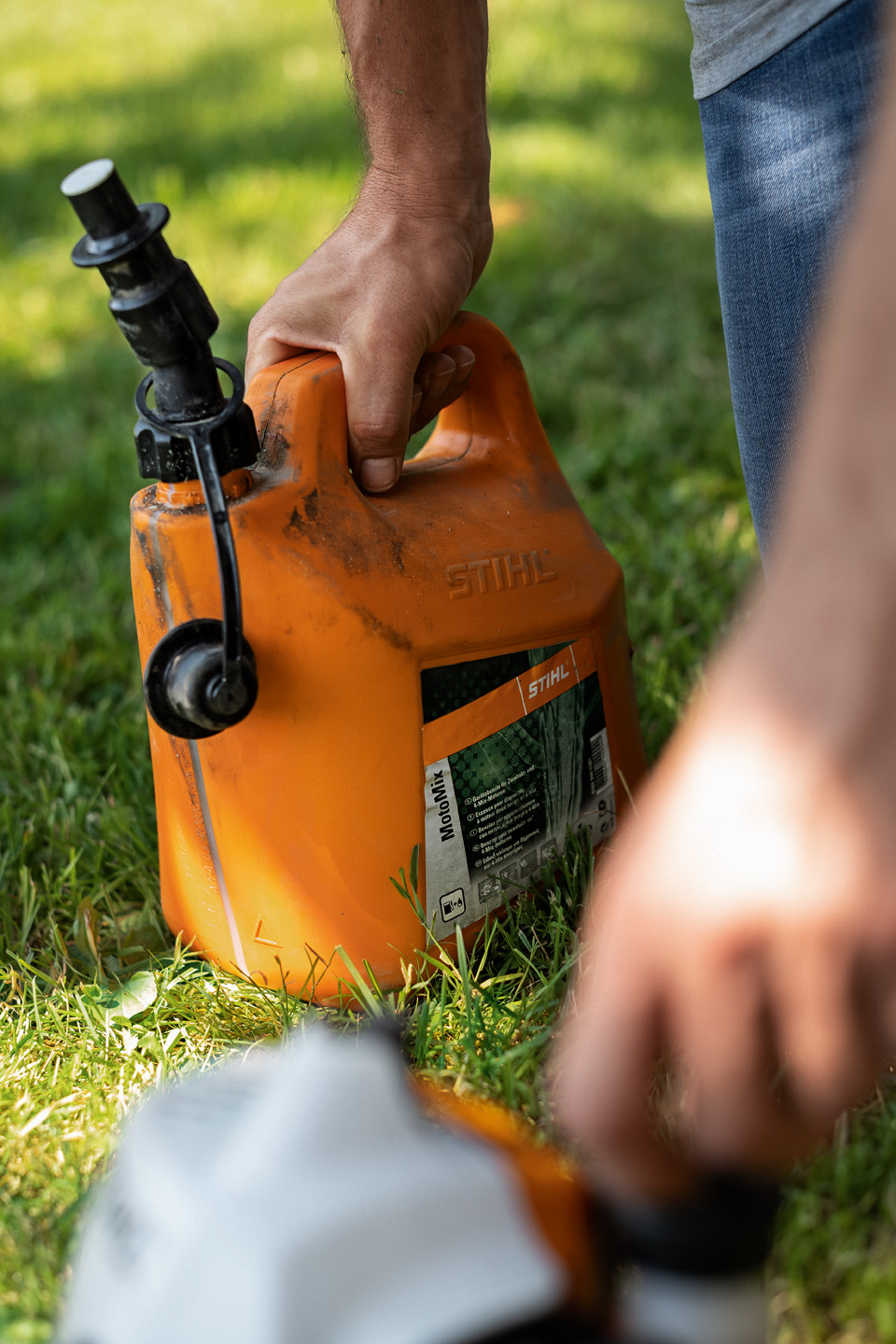 Recipiente sucio de una mezcla de combustible STIHL MotoMix sobre una superficie de hierba, con una mano abriendo el tapón de combustible de una máquina en primer plano